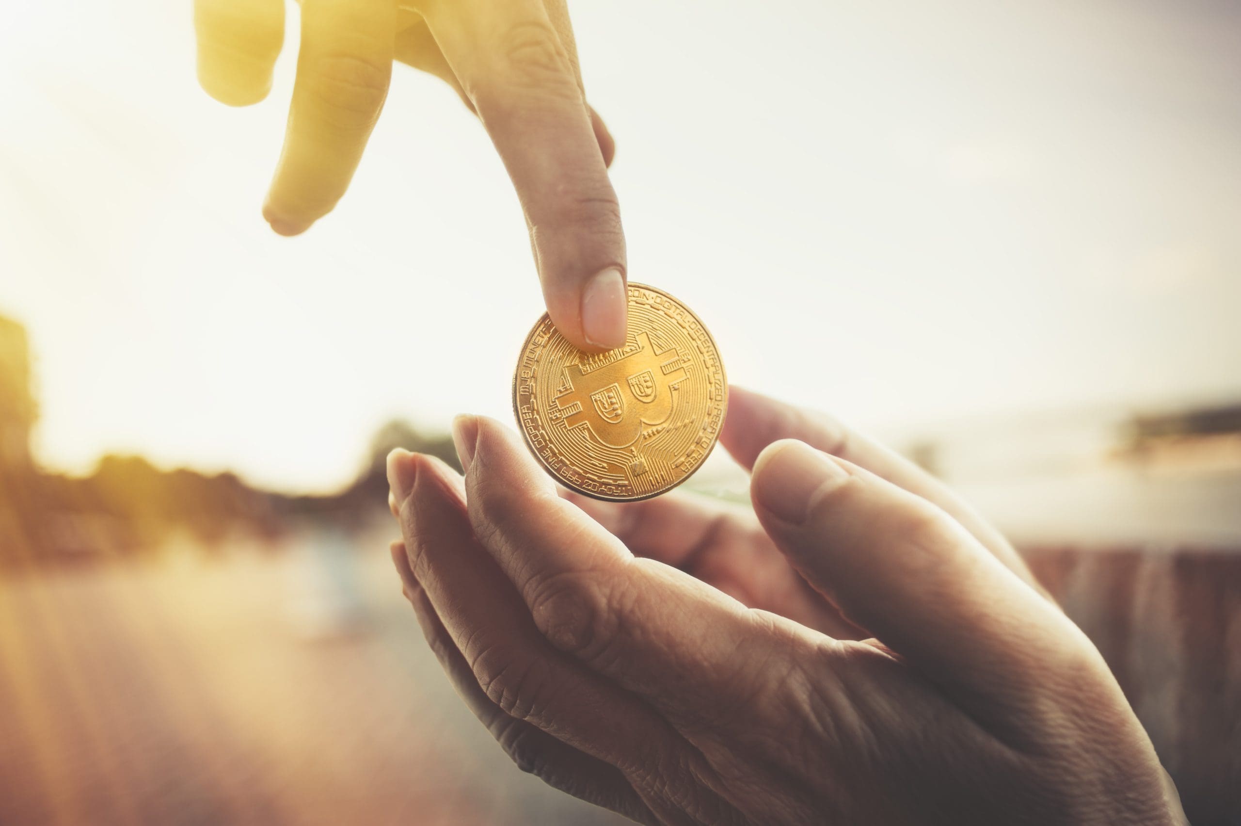 A sepia toned photo of a downward pointing pale-skinned hand holding a bitcoin dangles it above outstretched hands of another light-skinned person. There is a blurry background behind them.