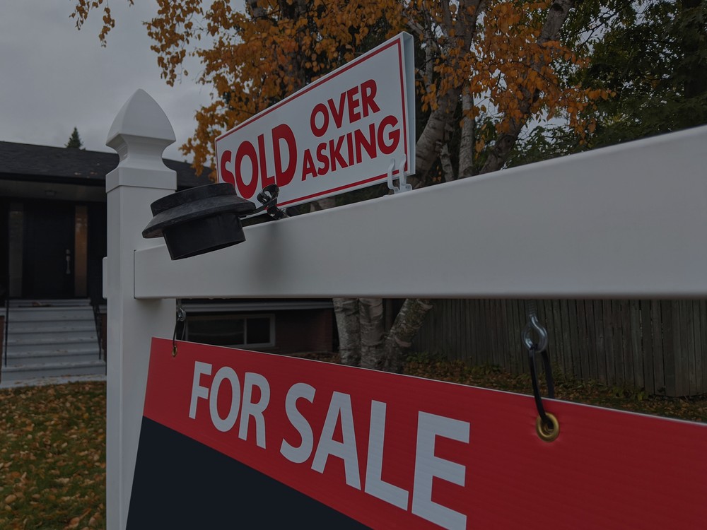 An image of a "For Sale" sign in the yard of a house, which includes a smaller noting that it "sold over asking" price.